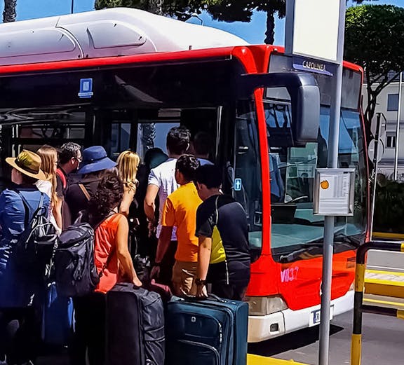 Passengers boarding a red bus with luggage at a sunny bus stop.