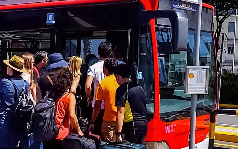 Passengers boarding a red bus with luggage at a sunny bus stop.