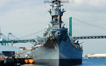 Battleship docked at port with bridge in background, self-guided experience available.