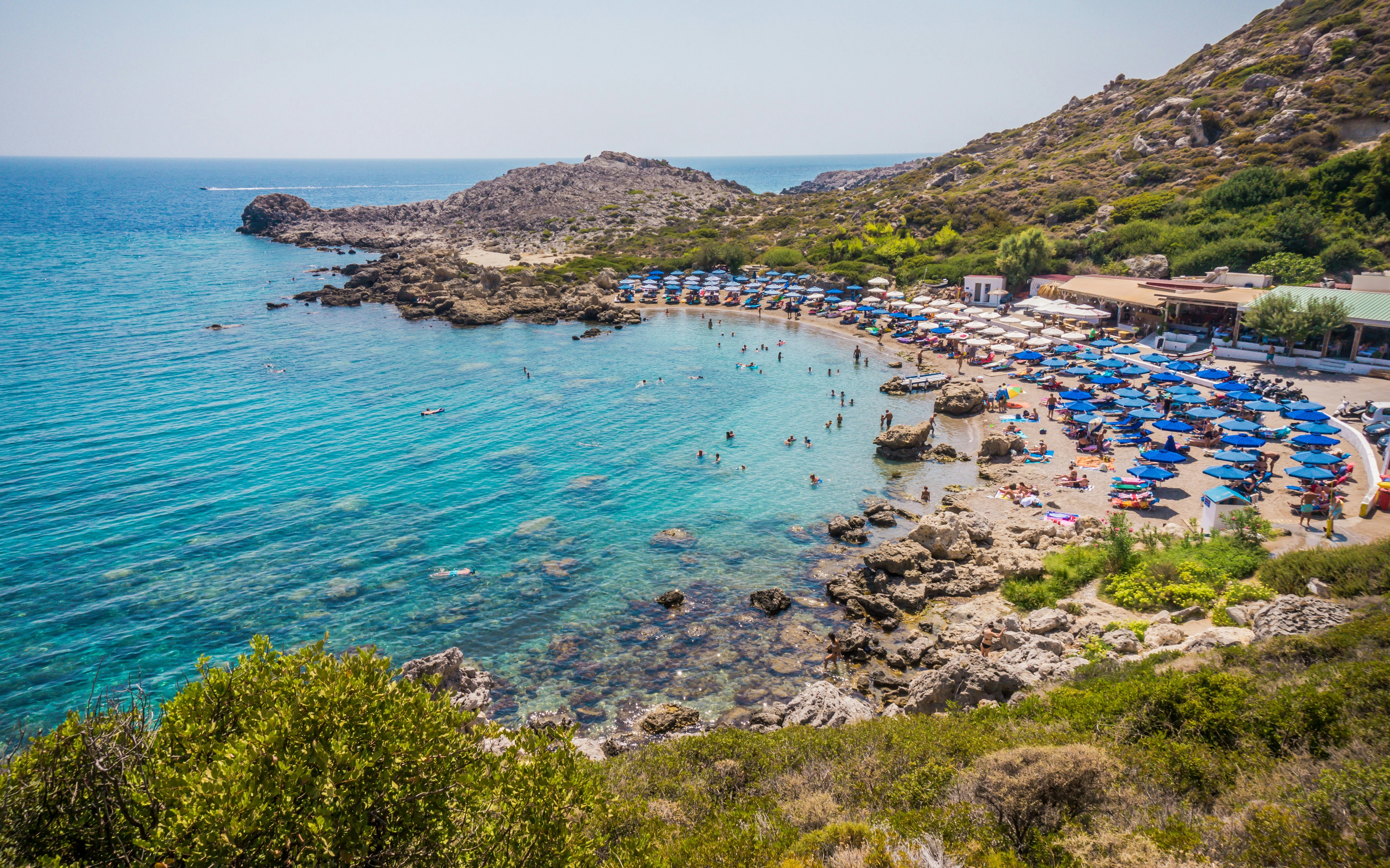 Ladiko Bay in Rhodes with beachgoers, blue umbrellas, and rocky shoreline.