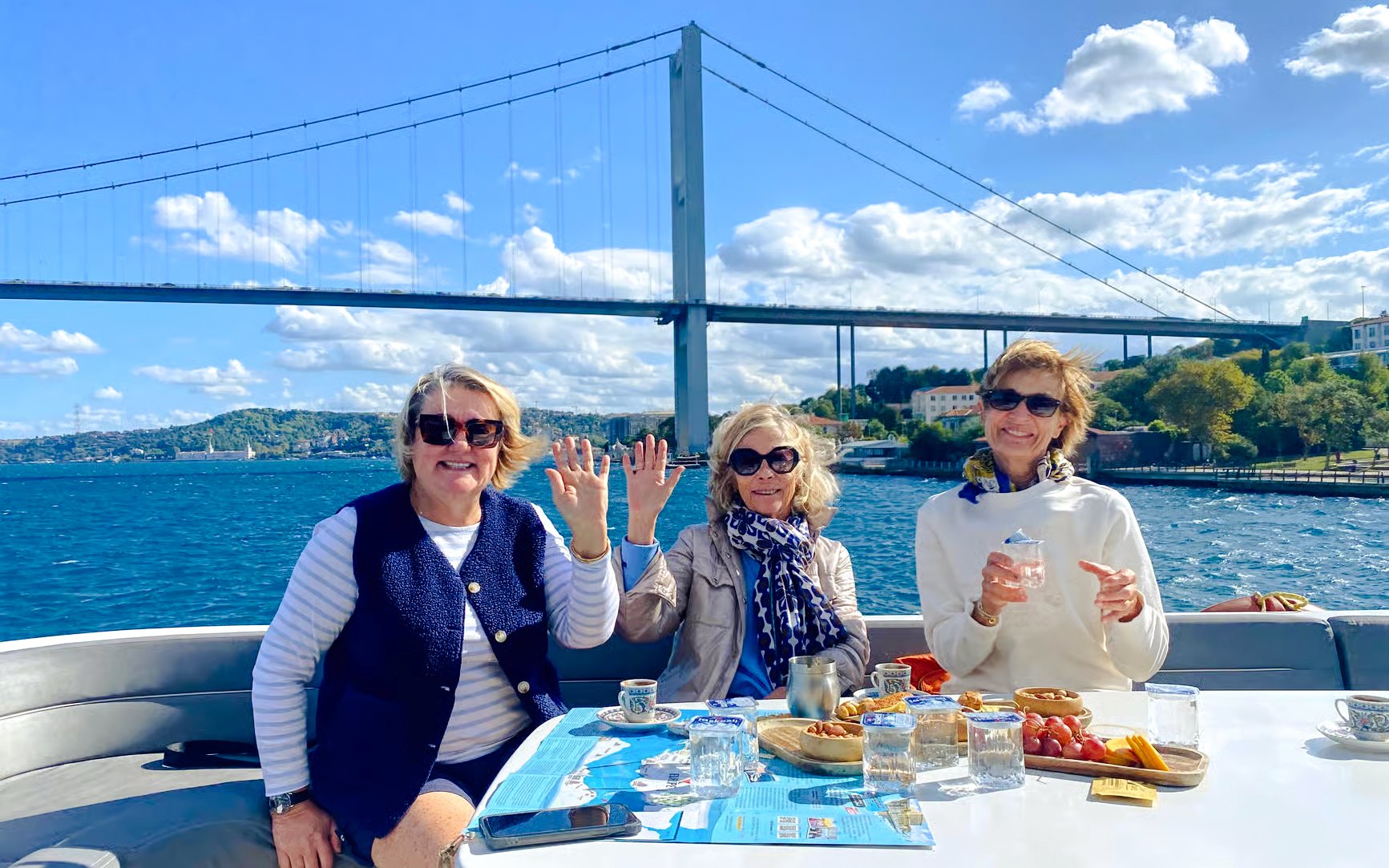 Tourists enjoying a Bosphorus yacht cruise with snacks and drinks, Istanbul bridge in background.