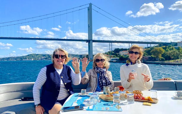 Tourists enjoying a Bosphorus yacht cruise with snacks and drinks, Istanbul bridge in background.