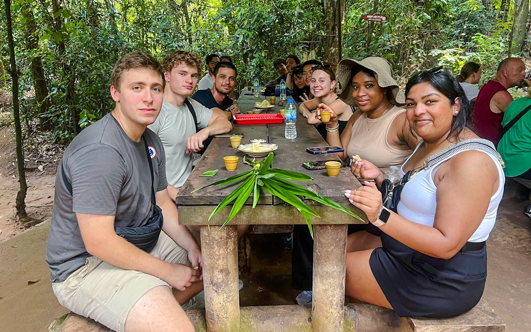 Tour group enjoying snacks at a wooden table in Cu Chi tunnels, Vietnam.