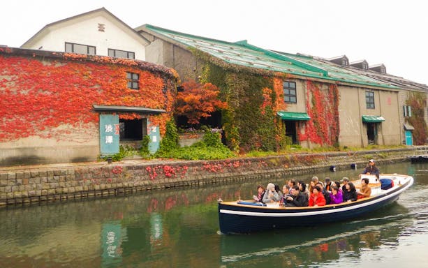 Boat tour on Otaru Canal with passengers, passing ivy-covered warehouses in Japan.