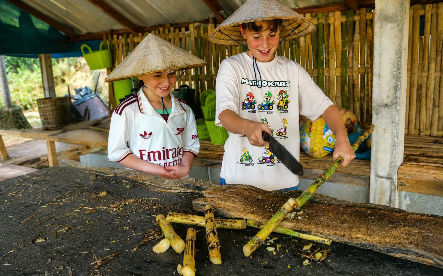 Two young visitors in straw hats chopping sugarcane in a bamboo shelter for elephant feeding.