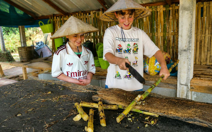 Two young visitors in straw hats chopping sugarcane in a bamboo shelter for elephant feeding.