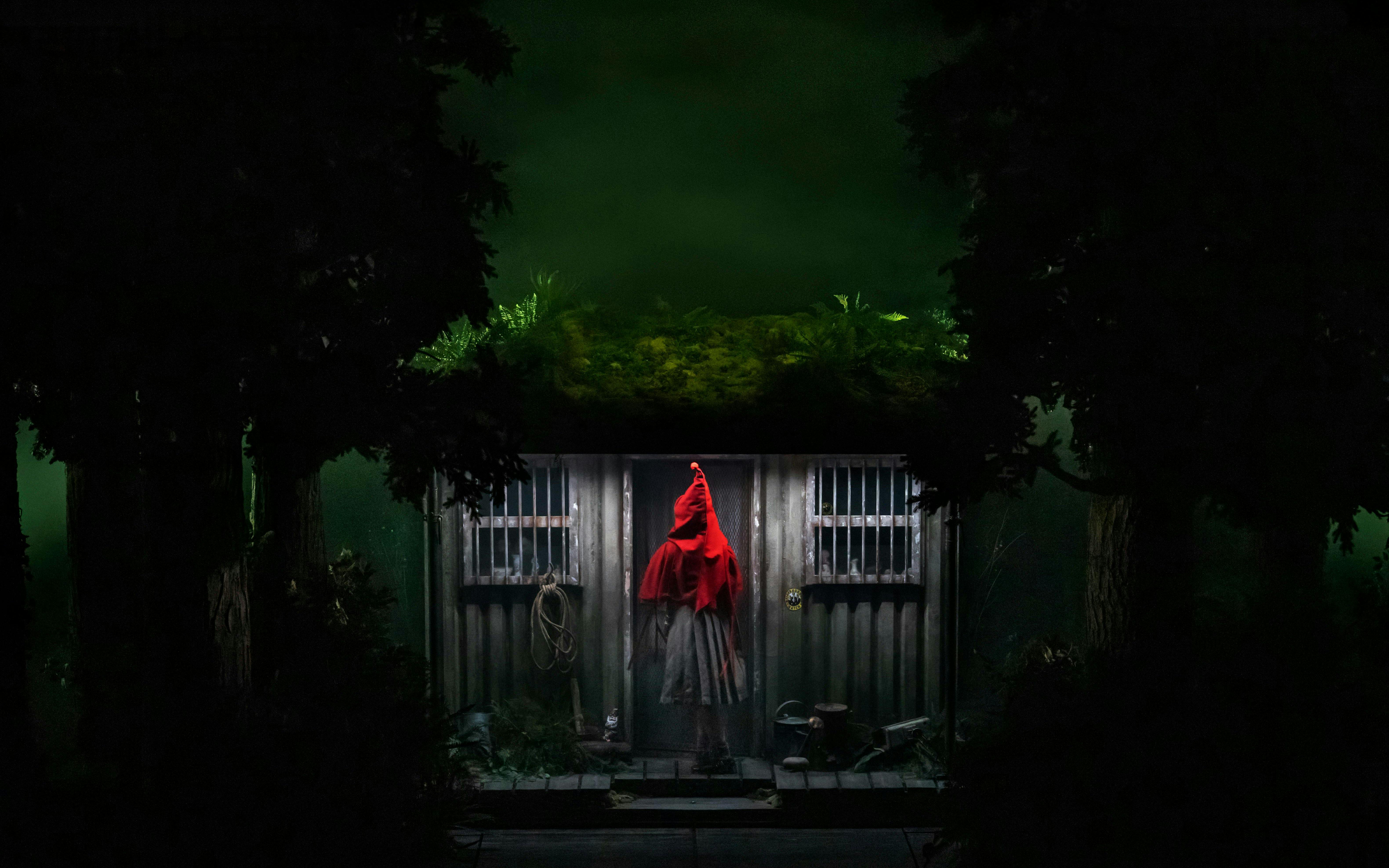Person in red cloak standing at a cabin door in a dark forest setting.