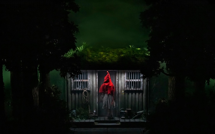 Person in red cloak standing at a cabin door in a dark forest setting.