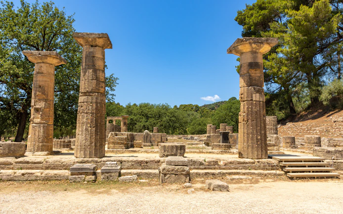 Ancient ruins of the Temple of Hera in Olympia, Greece, with stone columns and trees.