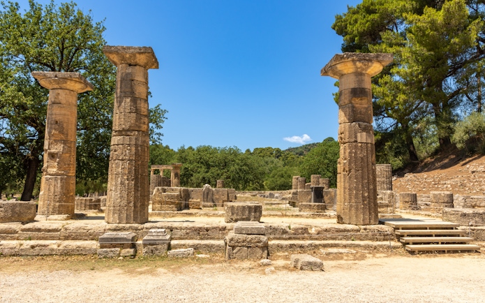 Ancient ruins of the Temple of Hera in Olympia, Greece, with stone columns and trees.