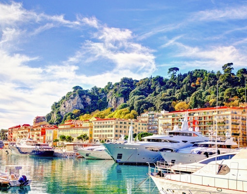 Der Hafen von Lympia in Nizza mit Yachten und bunten Gebäuden vor einem Hügel.