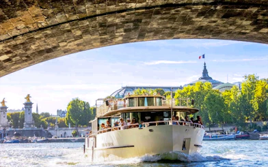 Seine River cruise boat passing under a bridge in Paris with the Grand Palais in the background.