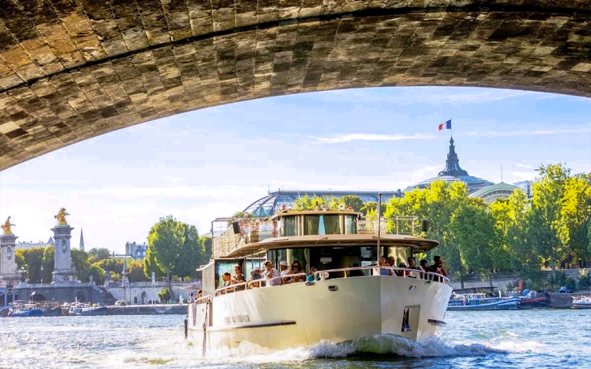 Seine River cruise boat passing under a bridge in Paris with the Grand Palais in the background.