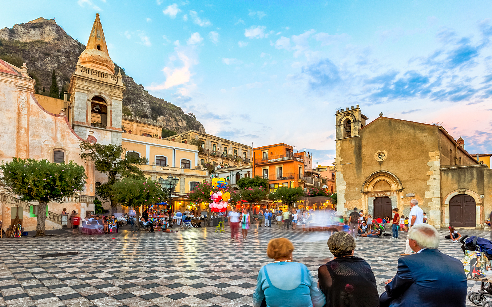 Piazza IX Aprile in Taormina with people, historic buildings, and mountain backdrop.