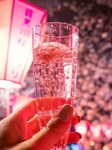 Hand holding a sparkling drink under cherry blossoms with lanterns in the background.
