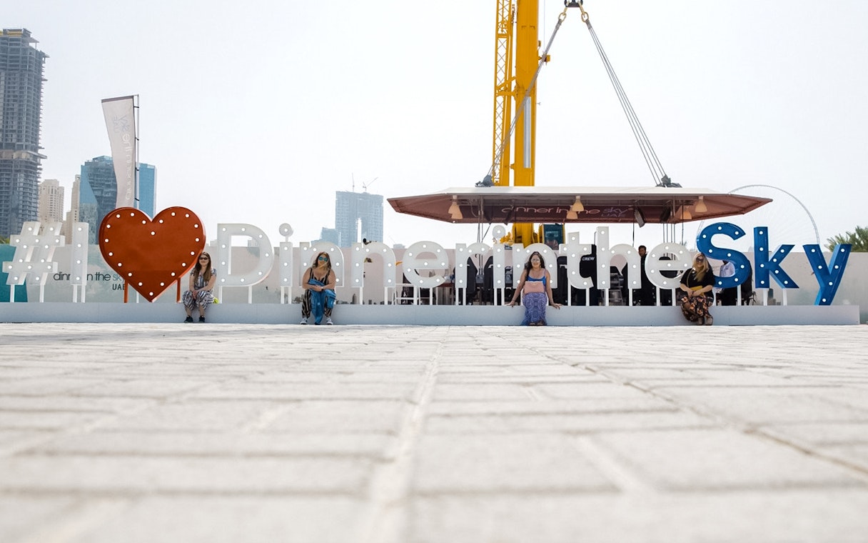 Dinner in the Sky sign with people seated, crane visible, city skyline in background.