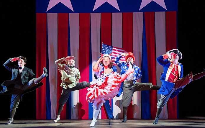 Performers in patriotic costumes dancing on stage with American flag backdrop.