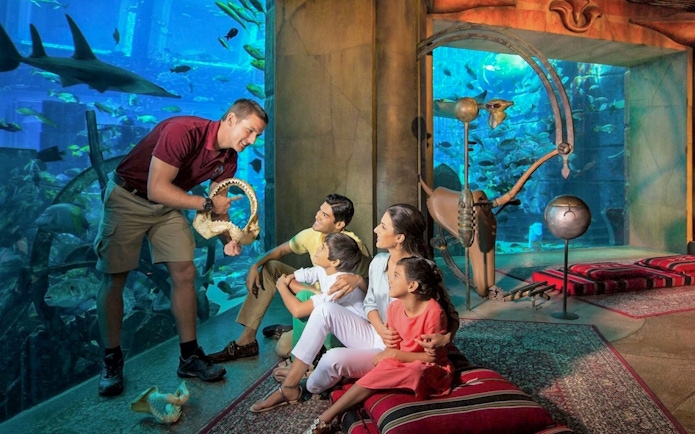 Family observing marine life at The Lost Chambers Aquarium, Dubai, with a guide showing a shark jaw.