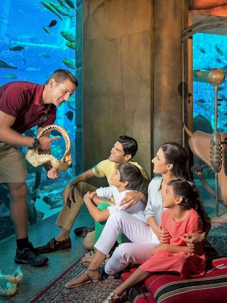 Family observing marine life at The Lost Chambers Aquarium, Dubai, with a guide showing a shark jaw.