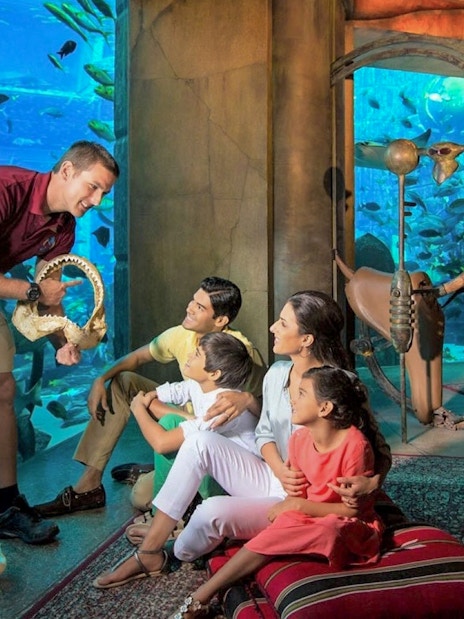 Family observing marine life at The Lost Chambers Aquarium, Dubai, with a guide showing a shark jaw.