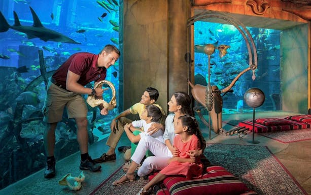 Family observing marine life at The Lost Chambers Aquarium, Dubai, with a guide showing a shark jaw.
