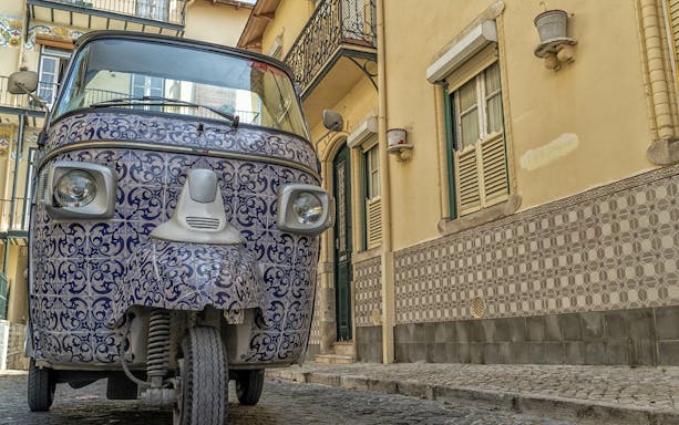 Electric tuk tuk with decorative tiles on a cobblestone street in a historic neighborhood.