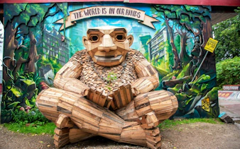 Wooden sculpture in Freetown Christiania, Copenhagen, with mural background and "The World is in Our Hands" sign.
