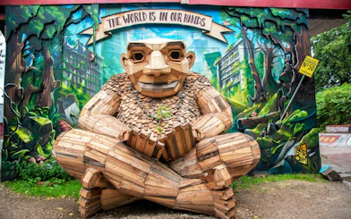 Wooden sculpture in Freetown Christiania, Copenhagen, with mural background and "The World is in Our Hands" sign.