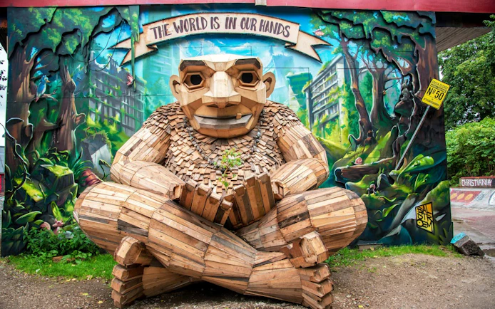 Wooden sculpture in Freetown Christiania, Copenhagen, with mural background and "The World is in Our Hands" sign.