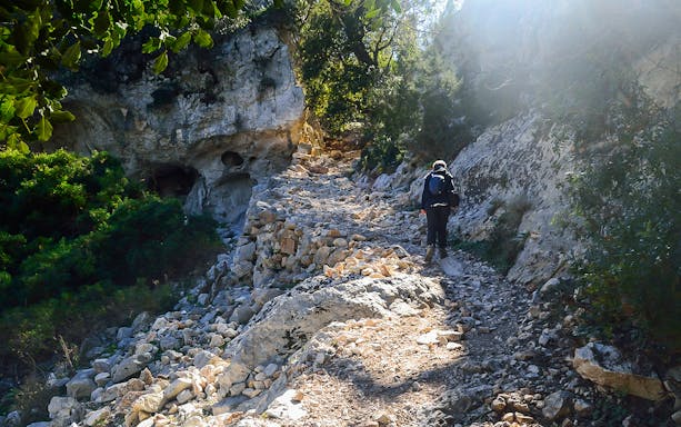 Hiker on rocky trail surrounded by cliffs and greenery in Cala Goloritzè, Sardinia.