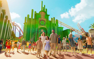 Tourists exploring Wizard of Oz precinct at Movie World with green buildings and roller coaster.