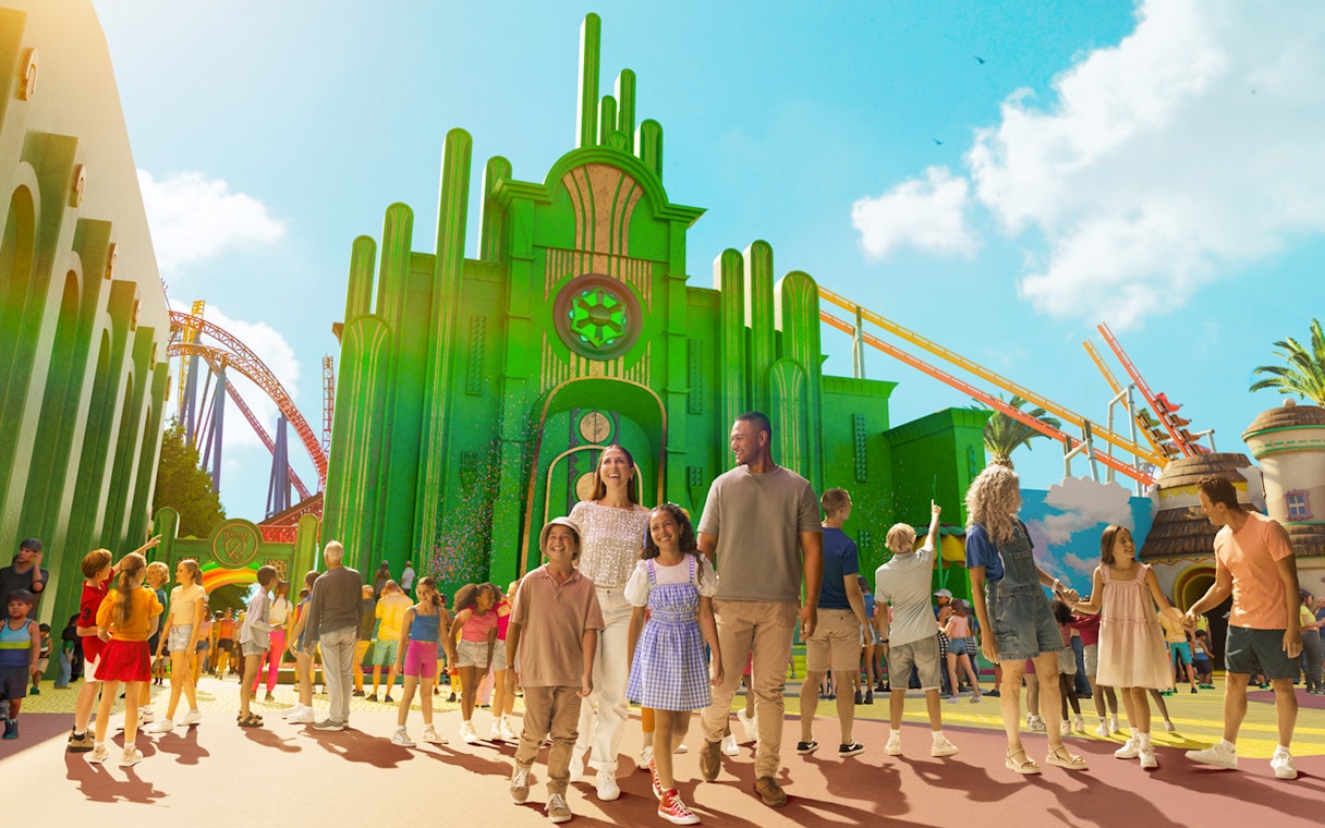 Tourists exploring Wizard of Oz precinct at Movie World with green buildings and roller coaster.
