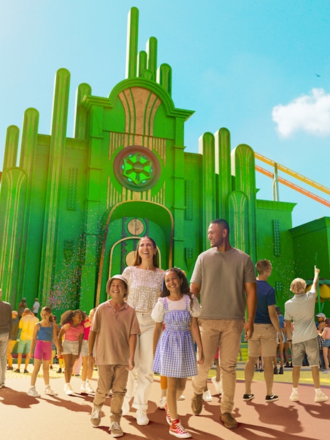 Tourists exploring Wizard of Oz precinct at Movie World with green buildings and roller coaster.