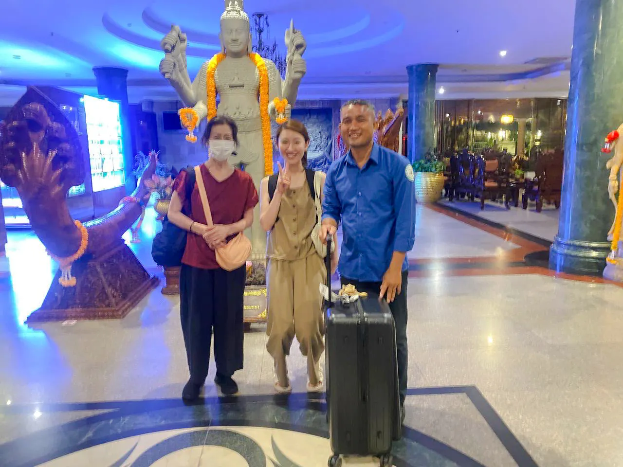 Travelers with luggage at Siem Reap Angkor Airport transfer service, standing by a statue.