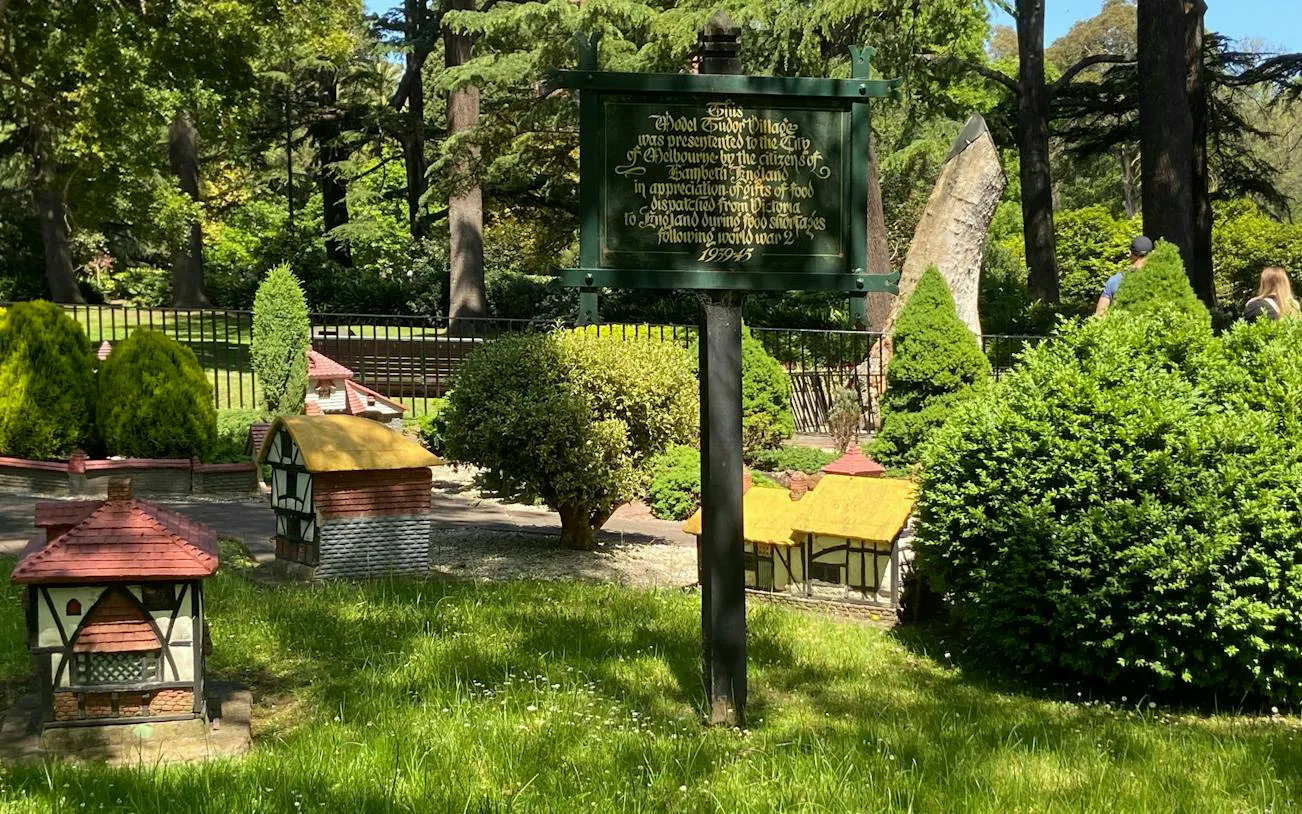 Miniature Tudor village in Fitzroy Gardens, Melbourne, surrounded by lush greenery and informational sign.