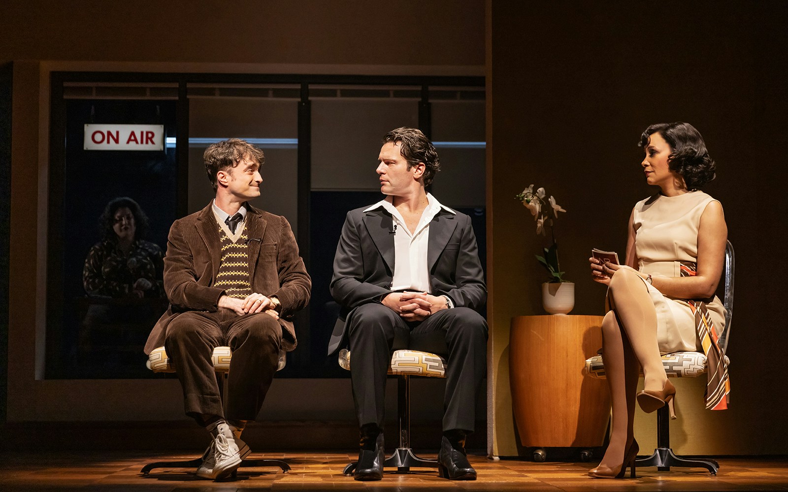 Three actors seated on stage during a scene from "Merrily We Roll Along" with an "On Air" sign in the background.