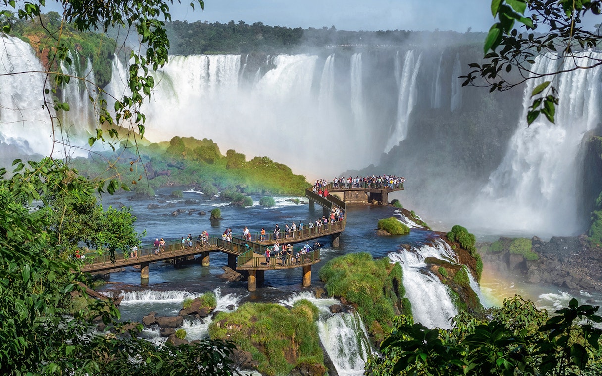 Tourists on a walkway at Iguazu Falls, surrounded by lush greenery and cascading water.