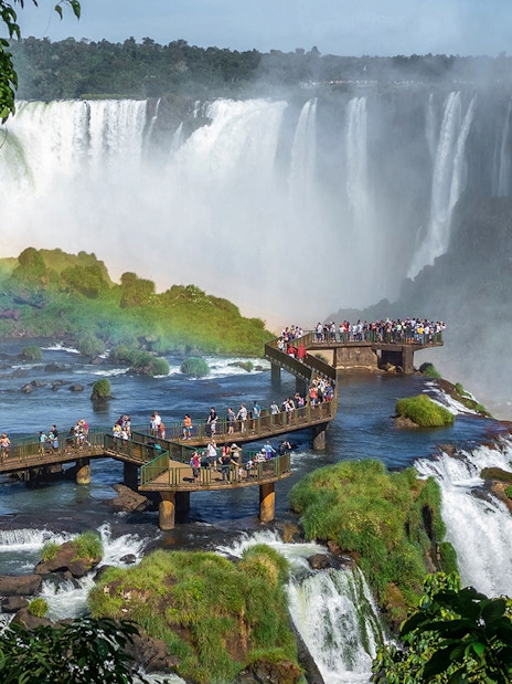 Tourists on a walkway at Iguazu Falls, surrounded by lush greenery and cascading water.