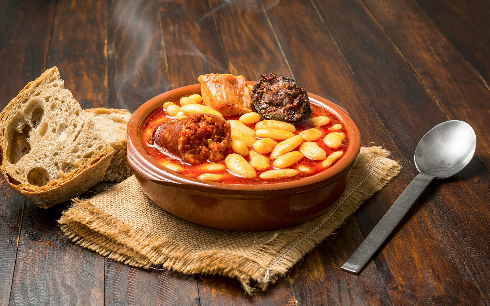 Steaming bowl of Spanish fabada with bread on a wooden table in Toledo restaurant.