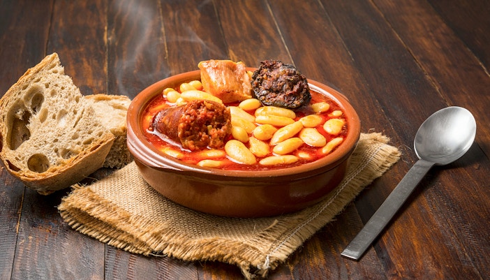 Steaming bowl of Spanish fabada with bread on a wooden table in Toledo restaurant.