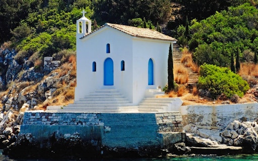 Chapel of Agios Andreas on rocky shore, Ithaca, Greece, surrounded by greenery.