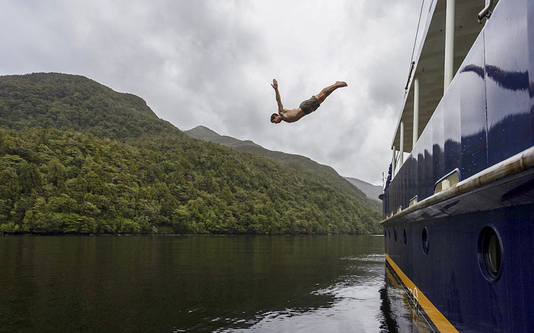 Person diving from a boat into Doubtful Sound, surrounded by lush hills, during an overnight cruise from Te Anau.