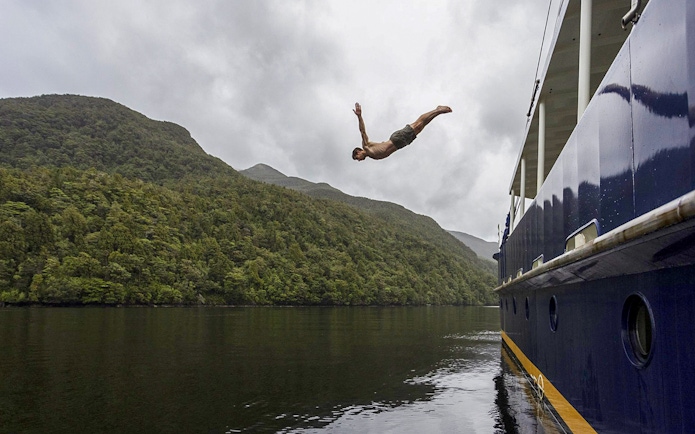 Person diving from a boat into Doubtful Sound, surrounded by lush hills, during an overnight cruise from Te Anau.