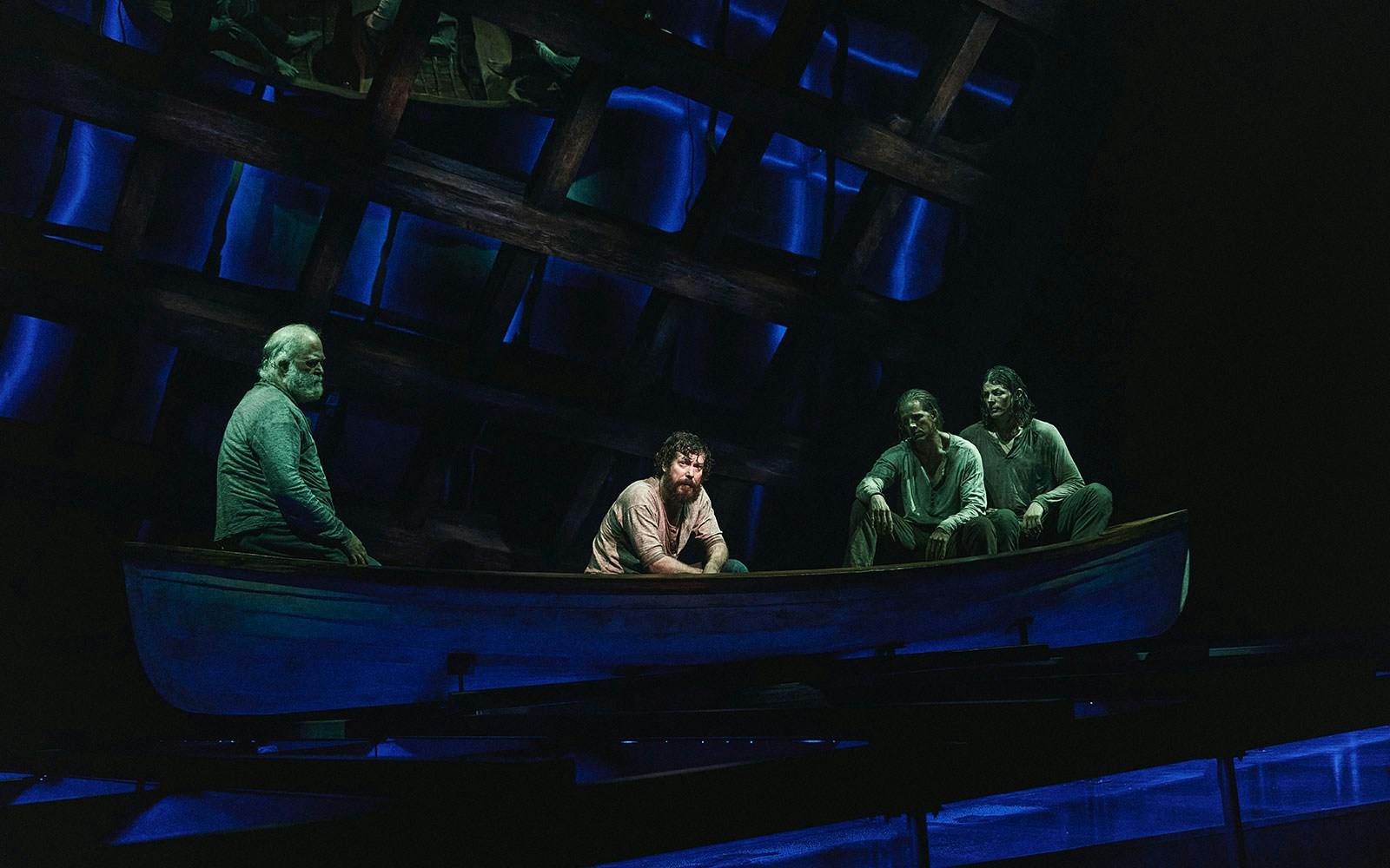 Men sitting in a boat on stage with a dramatic blue-lit backdrop, part of "Swept Away" performance.