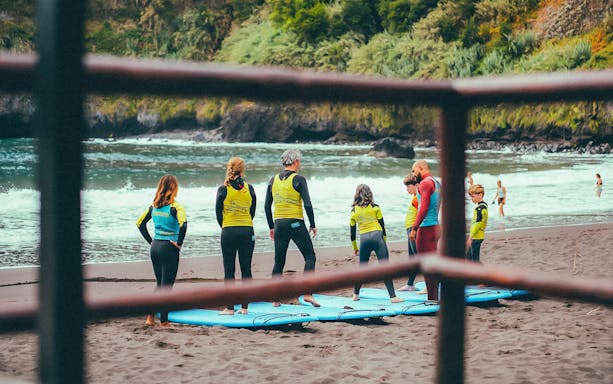 Family learning to surf on a beach with instructor, surrounded by lush cliffs.