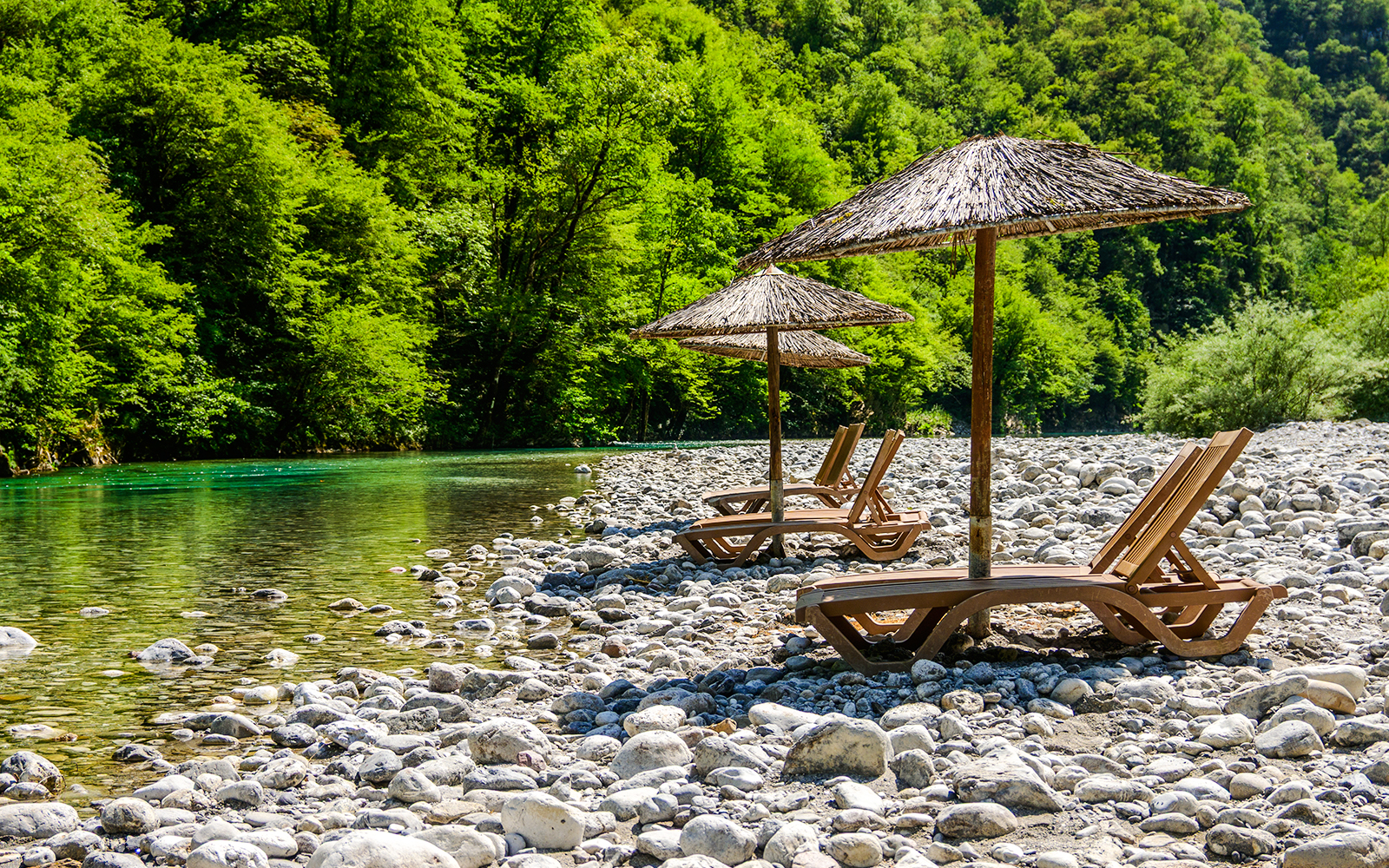 Sunbathing chairs with straw umbrellas by Shala River, surrounded by lush greenery.