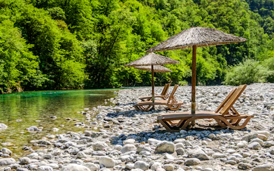 Sunbathing chairs with straw umbrellas by Shala River, surrounded by lush greenery.