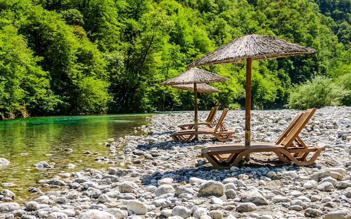 Sunbathing chairs with straw umbrellas by Shala River, surrounded by lush greenery.