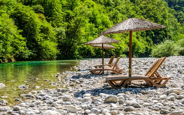 Sunbathing chairs with straw umbrellas by Shala River, surrounded by lush greenery.
