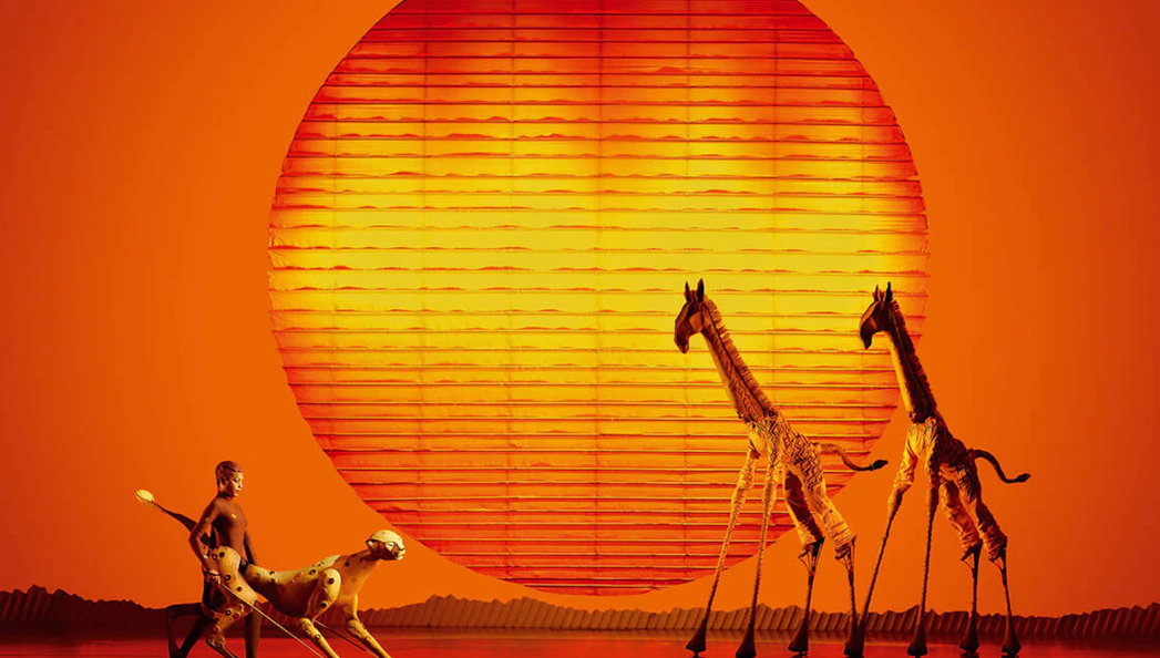 Stage performance of The Lion King with actors as animals against a sunset backdrop.